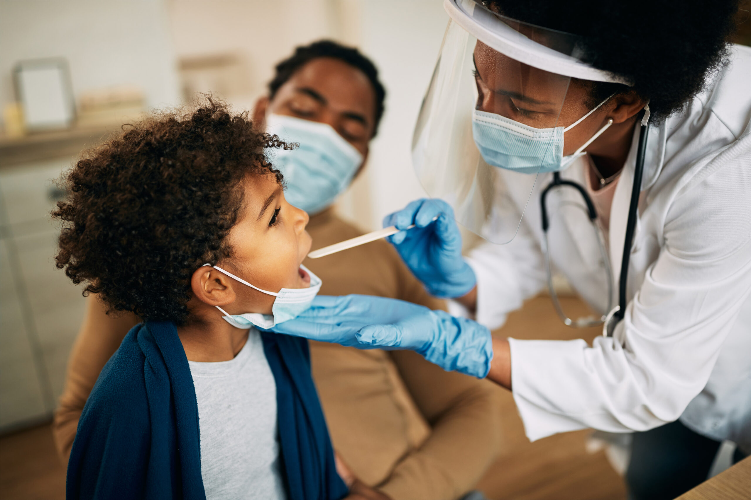 Family doctor examining throat of a small black boy while visiting him at home during coronavirus pandemic.